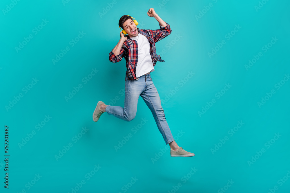 Full length photo of funky excited man dressed plaid shirt jumping high rising fists enjoying music isolated turquoise color background