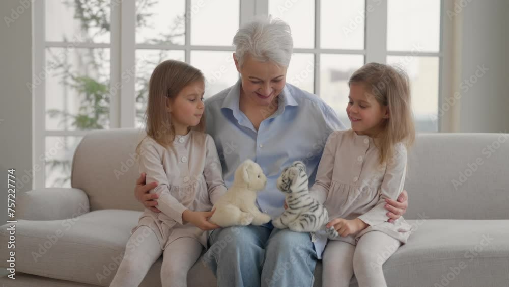 Grandmother with granddaughters resting. Happy relaxed older adult granny embracing playing with toys with children twins grandchildren leisure rest on couch at home. Family love, generation concept. Grandmother with granddaughters resting. Happy relaxed older adult granny embracing playing with toys with children twins grandchildren leisure rest on couch at home. Family love, generation concept.