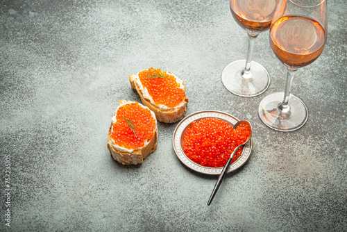 Small metal plate with red salmon caviar, two caviar toasts canape, two glasses of champagne top view on grey concrete background, festive luxury delicacy and appetizer.