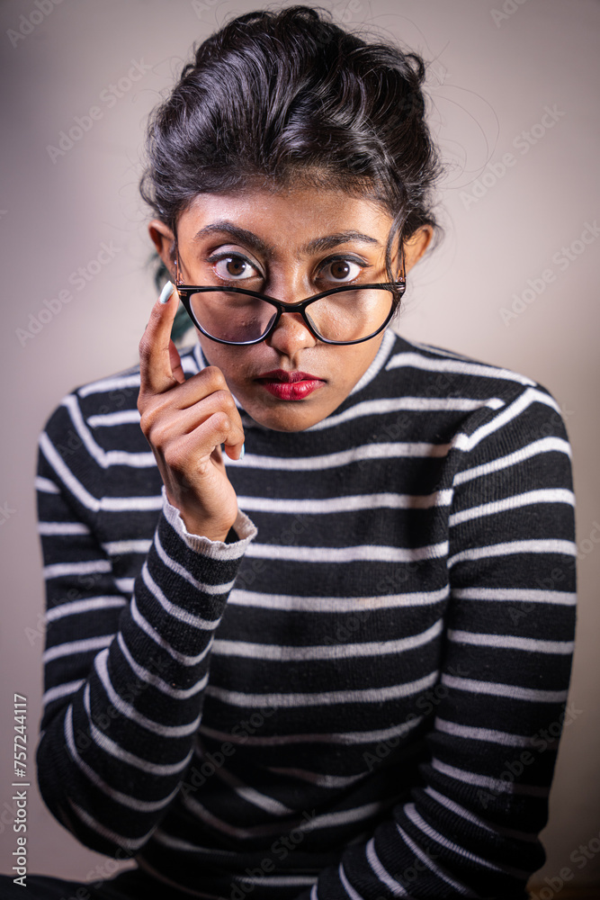 A woman with glasses is sitting and looking at the camera. She is wearing a striped shirt and has red lipstick on. Concept of curiosity and intrigue