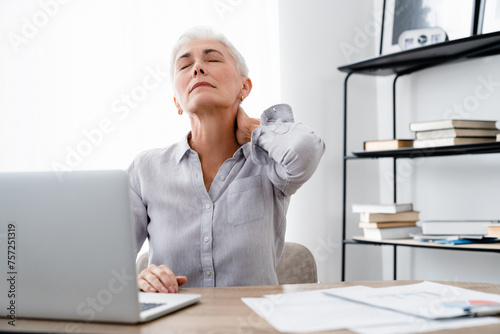 Serious Caucasian middle-aged businesswoman suffering from neck back pain in office. Mature manager overworked using laptop, rubbing painful muscles photo