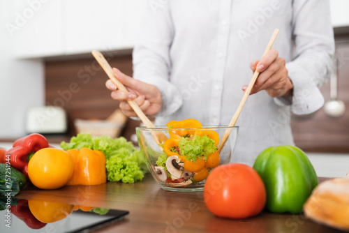 Cropped image of woman mixing with spoons vegetable salad in bowl at home kitchen. Middle-aged housewife preparing cooking making vegan vegetarian salad indoors