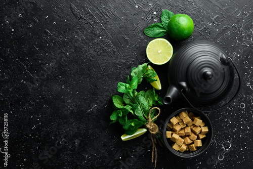 Mint tea with lime: fresh green limes, mint, brown sugar and a kettle of boiling water. On a black stone background. Top view.