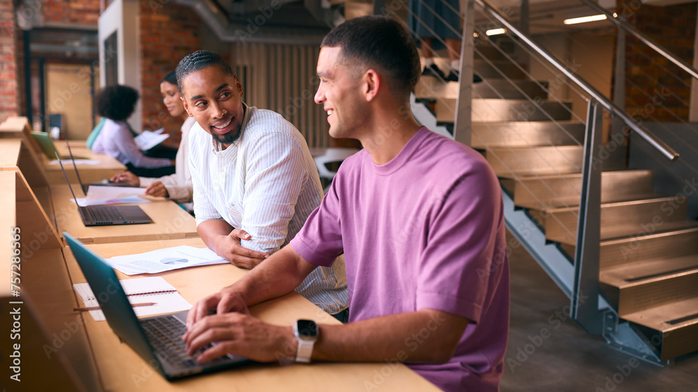 Two Businessmen With Laptop Meeting In Office Discussing Documents With Colleagues In Background Two Businessmen With Laptop Meeting In Office Discussing Documents With Colleagues In Background