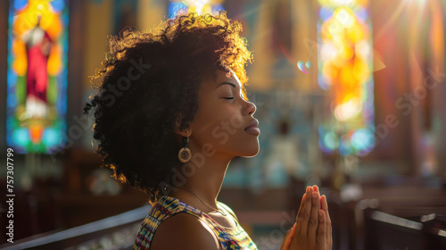 A serene young black woman praying in a sunlit church with stained glass windows in the background.