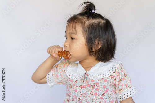 A happy little girl stands interestingly eating crispy fried chicken thighs. , The bright and cute little girl and the delicious fried chicken legs were happy to eat the delicious food. photo