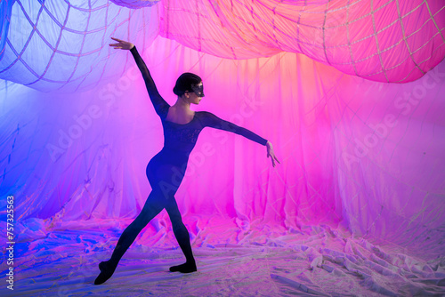 Ballerina dancer in a room with curtains made of thin airy fabric, illuminated by blue and red spotlights.