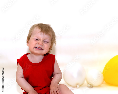 One-year-old toddler with blonde hair in red bodysuit sitting and smiling on white background with inflatable balloons. Wonderful kid sat down to rest. Baby posing isolated over light background