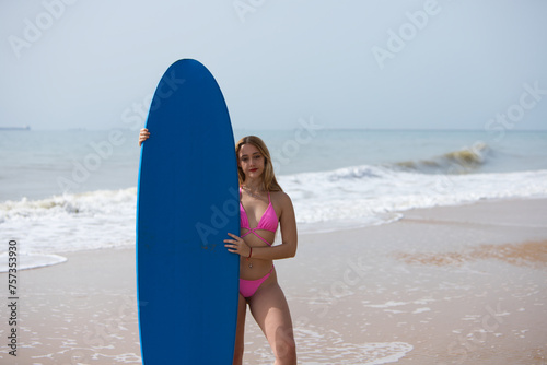 Young and beautiful blonde surfer woman in pink bikini and blue surfboard. The girl enjoys her holidays on the beach to practice her favourite sport. Travel and holiday concept