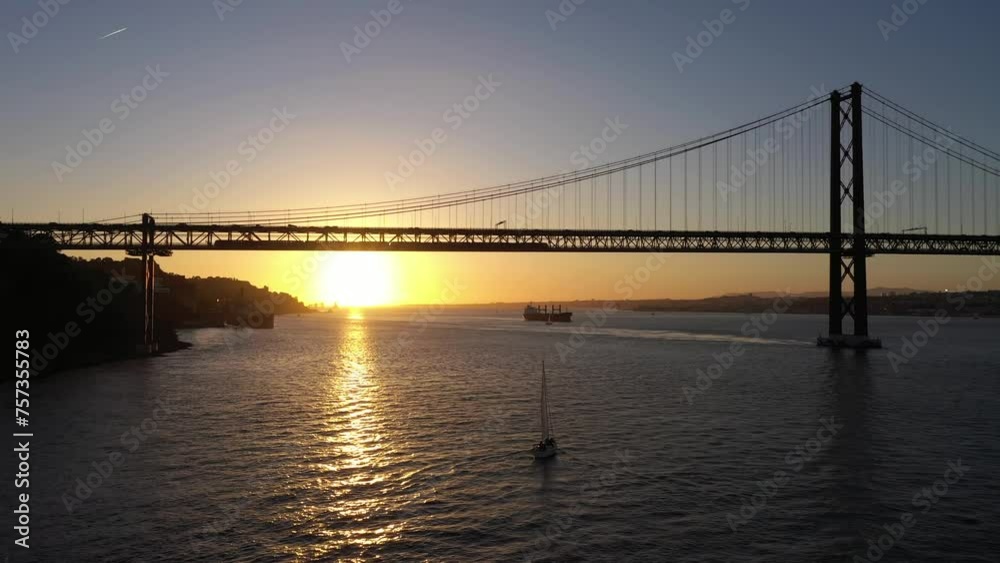 Silhouette of Ponte 25 de Abril Suspension Bridge at Sunset and Sailing Yacht. 25th April Bridge. Portugal. Aerial View. Drone Moves Forward and Upwards