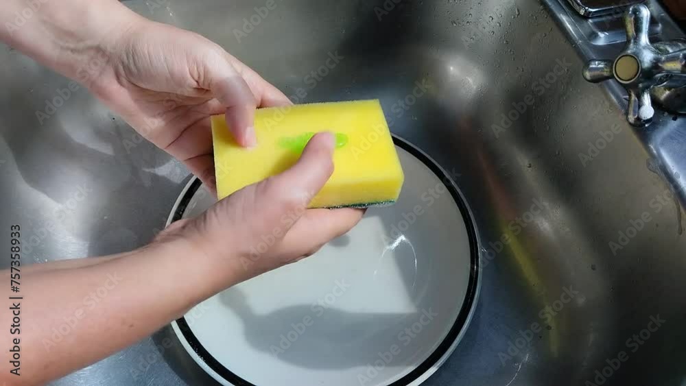 Unas manos de mujer lavando un plato de color blanco con una esponja, jabón liquido y agua en un fregadero Unas manos de mujer lavando un plato de color blanco con una esponja, jabón liquido y agua en un fregadero