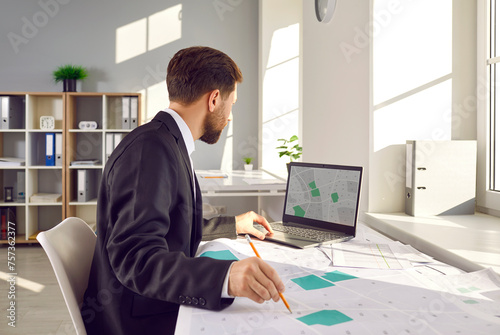 Man who is a professional corporate cadastral surveyor works with cartographic cadastre city maps, studies plot boundaries and numbers, uses digital graphic urban structure design plans on notebook PC photo