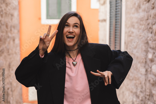 Cool brunette woman in black jacket, pink long sleeves top and heart shape necklace shows sign v, peace and friendship, open mouth and posing outside. Happy woman walk and posing outdoor in the city. 