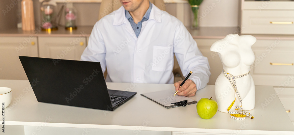 Online consultation with nutritionist. Wellness clinic web line. Young nutritionist working on laptop, writing diet plan for patient, consulting online. Apple, tape measure, body sculpture on desk Online consultation with nutritionist. Wellness clinic web line. Young nutritionist working on laptop, writing diet plan for patient, consulting online. Apple, tape measure, body sculpture on desk