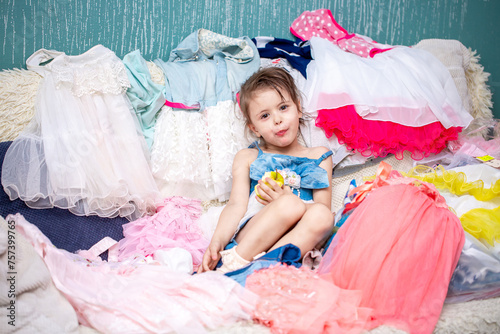 Little girl chooses clothes while sitting on the couch. Different outfits to choose from in the dresser. Shopaholic bought herself a lot of things.