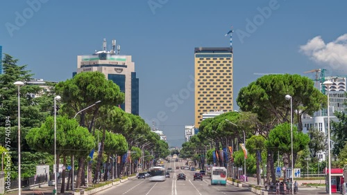 Traffic on the Deshmoret e Kombit Boulevard in Tirana timelapse. Main street in Albanian capital surrounded by green trees and skyscrapers. Cars moving and stops on intersections with a traffic light photo