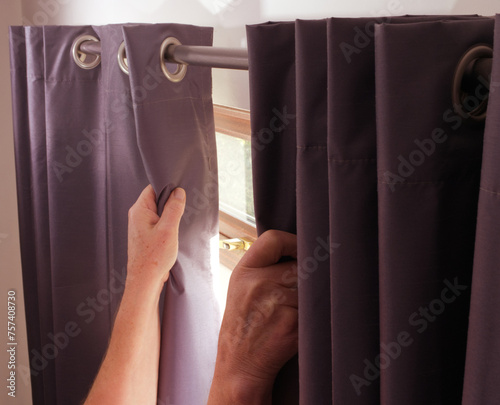 Fototapeta Closeup of hands opening or closing a pair of fabric curtains, attached on rings and sliding along a pole, in front of a room window