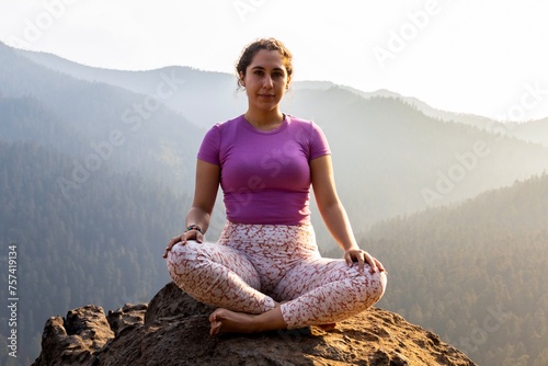 A woman in a yoga pose sits tranquilly atop a mountain, as the setting sun casts a warm glow over the forested landscape