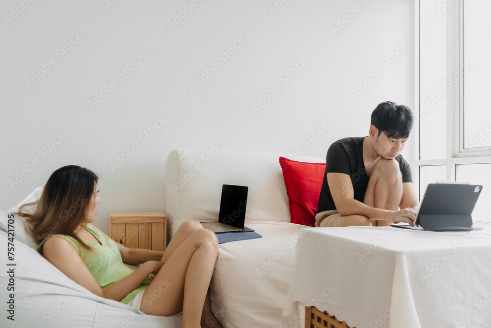 Asian Thai couple using laptop, Both man and woman busy working on notebook, lover freelancer sitting on couch in room apartment together.