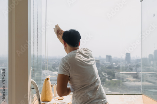 Backside view of asian Thai man wiping window glass in room apartment with city view, keep glass sparking clean, house husband and housework concept.