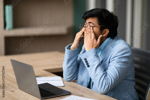 Stressed Indian businessman at laptop rubs tired eyes at office