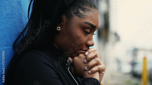 One pensive young black woman pondering decision with hands clenched together standing outside with thoughtful gaze struggling with dilemma in in urban setting