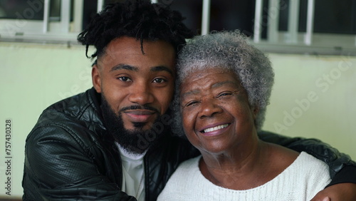 African American young grandson embracing his elderly gray hair grandmother portrait faces looking at camera in genuine loving moment, tender arm around shoulder, intergenerational bond