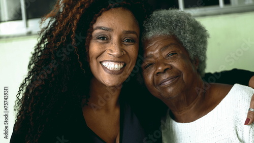 Tender loving moment of African American adult daughter kissing elderly 80s senior mother in the forehead and posing close-up faces smiling at camera