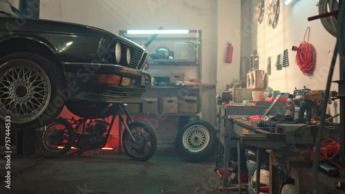 Black car on auto lift in garage for repair and maintenance service. Home suburban car garage interior with metal shelf, tools and equipment stuff storage warehouse on wall indoors.