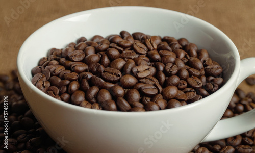 A coffee cup full of roasted coffee beans. Grains scattered around the cup. Close up.