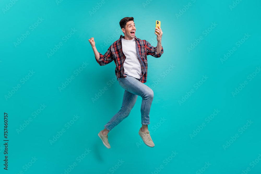 Full length photo of cheerful lucky guy wear checkered shirt jumping high winning game modern device isolated teal color background Full length photo of cheerful lucky guy wear checkered shirt jumping high winning game modern device isolated teal color background