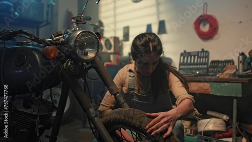 Beautiful female mechanic inspecting vintage motorbike condition in garage. Young woman biker tuning motorcycle in workshop. Retro motorcycle parts. Girl biker lifestyle concept.