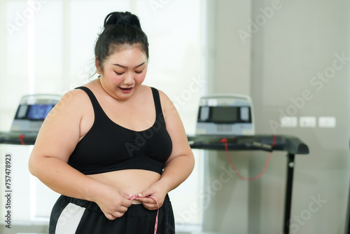 Chubby Asian woman exercise in gym, Joyful attire proudly measures her waist, showcasing positive results with a treadmill backdrop. fitness gear smiling while measuring waistline