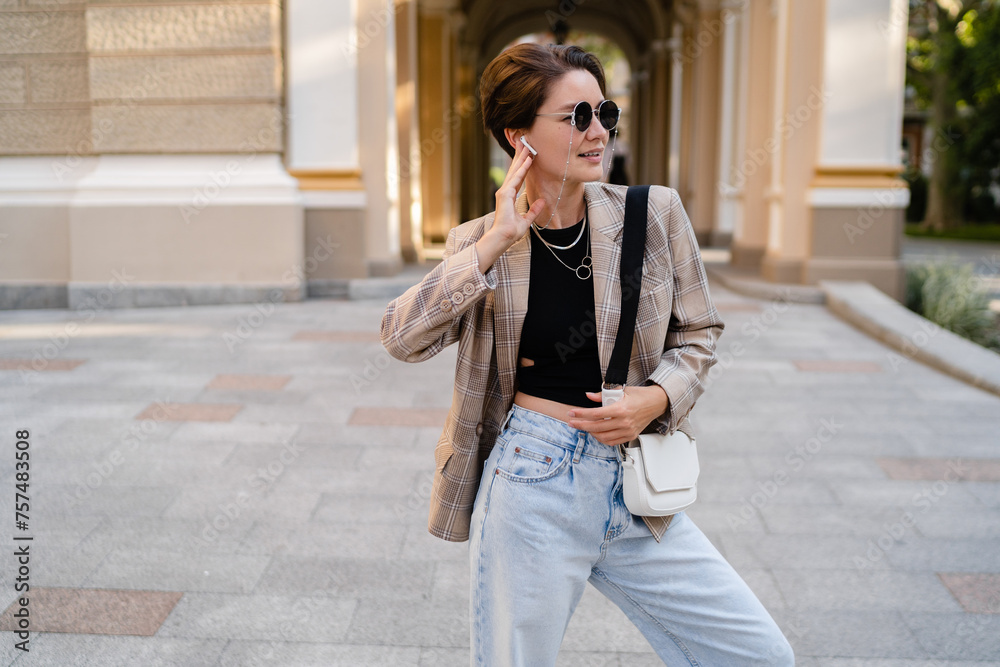 Fototapeta premium stylish woman with short haircut walking in street with coffee wearing beige jacket, jeans and sunglasses accessories