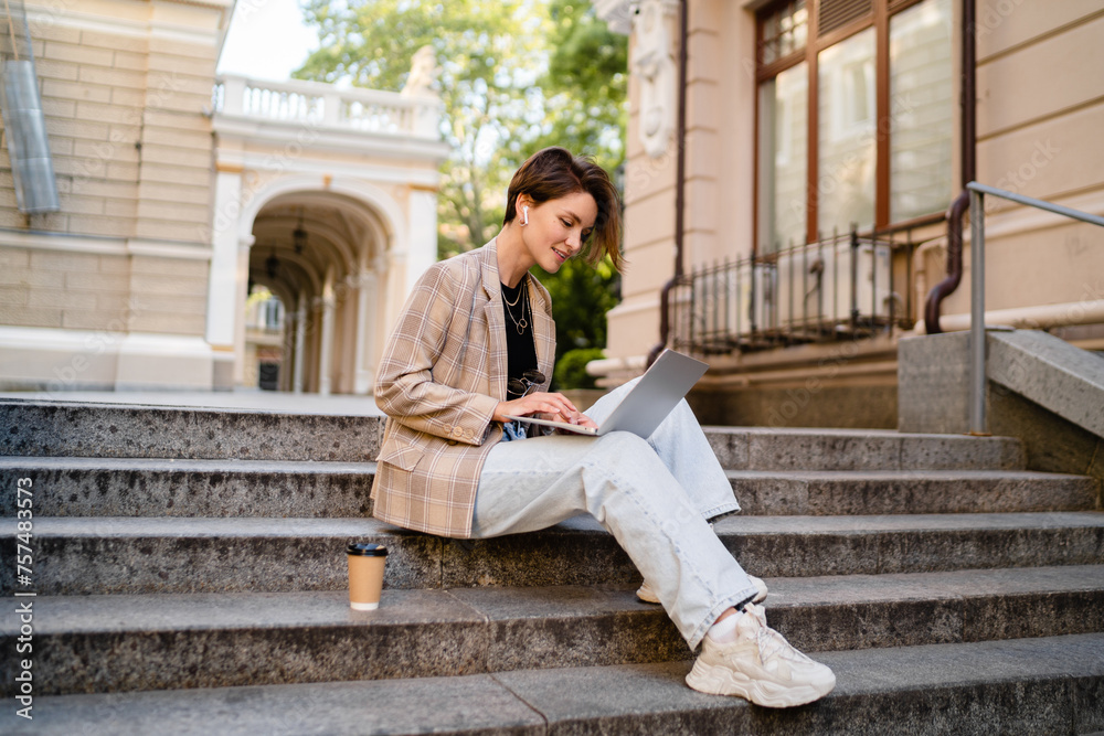 stylish woman with short haircut sitting in street wearing beige jacket, jeans working on laptop