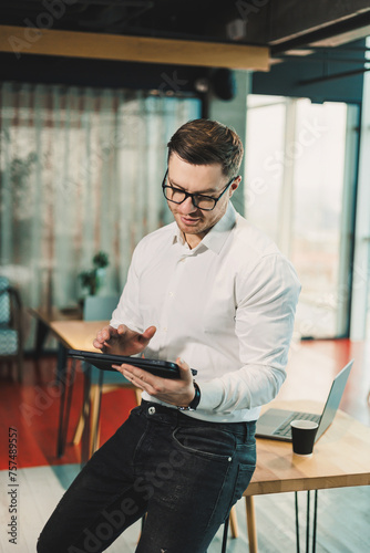 A young successful man in glasses and a white shirt is working on a tablet on a project in a modern workspace. Male businessman manager works in the office.