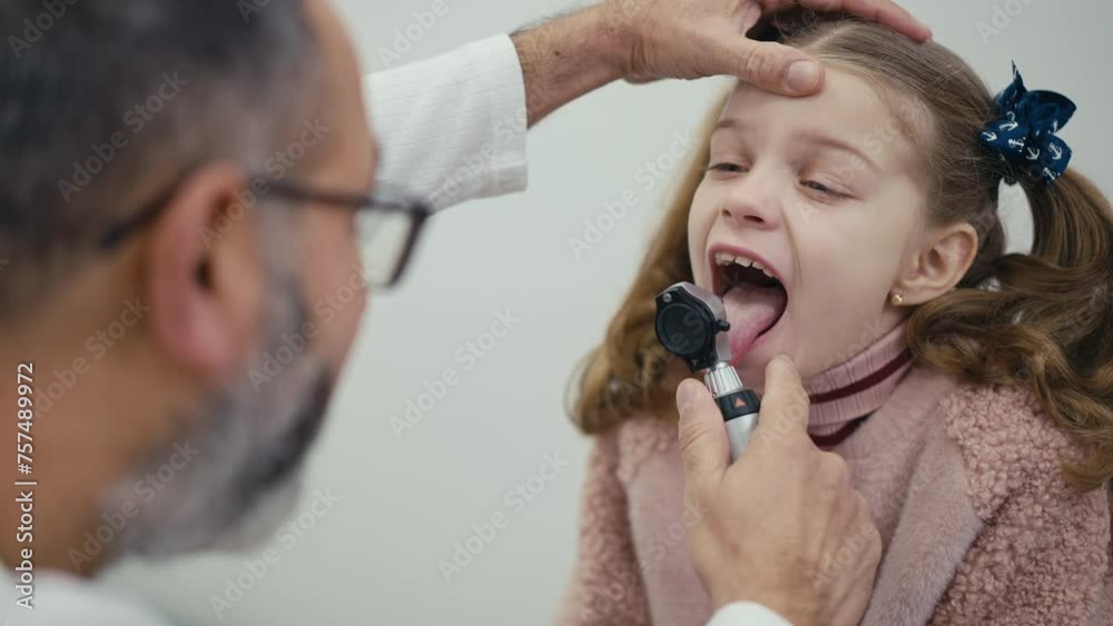 Shooting over the shoulder. A pediatrician performs a visual examination of the throat of a patient sitting on an examination couch in a modern clinic. A man doctor examines a little girl. The girl Shooting over the shoulder. A pediatrician performs a visual examination of the throat of a patient sitting on an examination couch in a modern clinic. A man doctor examines a little girl. The girl