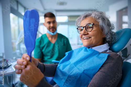 Satisfied senior woman using mirror while checking her teeth after procedure at dentist's office. photo