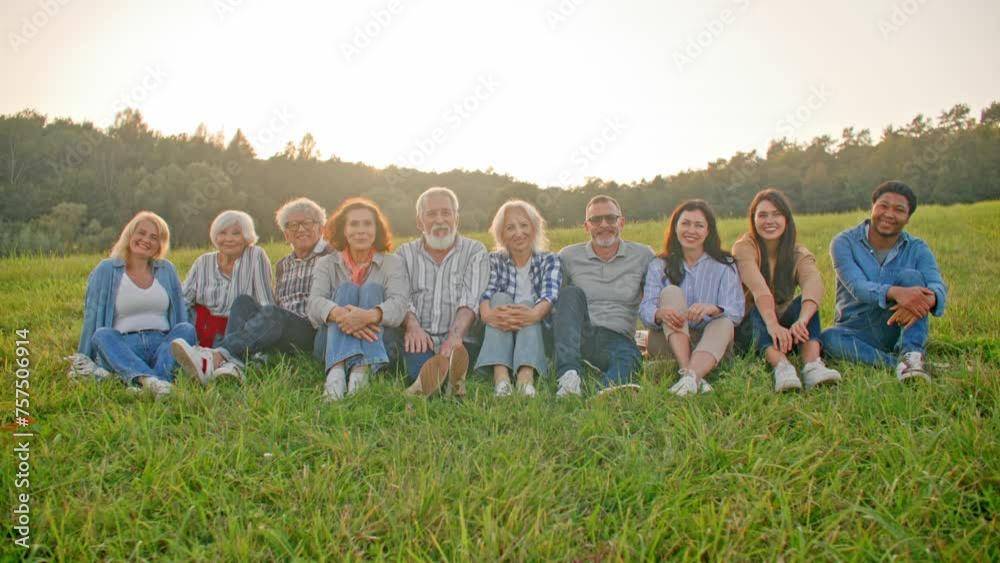 Group of multi-ethnic colleagues sitting on green grass next to each other. Looking directly at camera and smiling. Posing for perfect camera shot. Large forest behind in background. Group of multi-ethnic colleagues sitting on green grass next to each other. Looking directly at camera and smiling. Posing for perfect camera shot. Large forest behind in background.