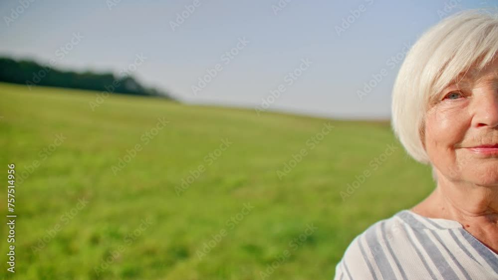 Portrait of older Caucasian woman standing in middle of field. Half of face visible, as person begins smiling. On side visible hill with green grass and forest in distance. Nature concept. Portrait of older Caucasian woman standing in middle of field. Half of face visible, as person begins smiling. On side visible hill with green grass and forest in distance. Nature concept.
