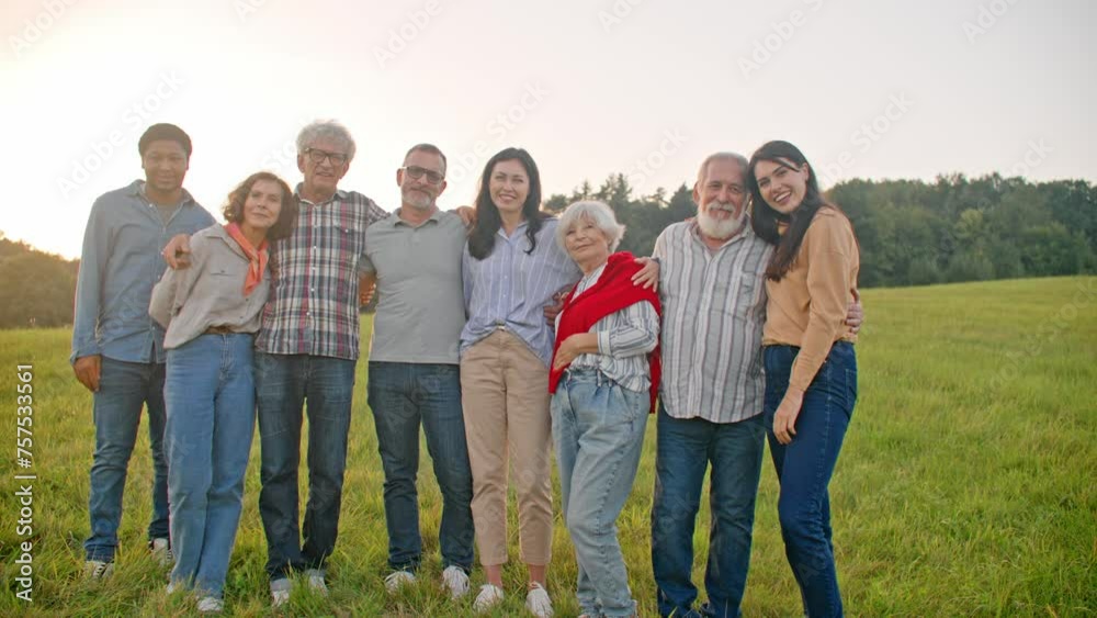 Multi-ethnic group of people hugging each other and smiling while looking directly at camera. Large field around friends. Team standing on green hill. Bright sunlight in background. Multi-ethnic group of people hugging each other and smiling while looking directly at camera. Large field around friends. Team standing on green hill. Bright sunlight in background.