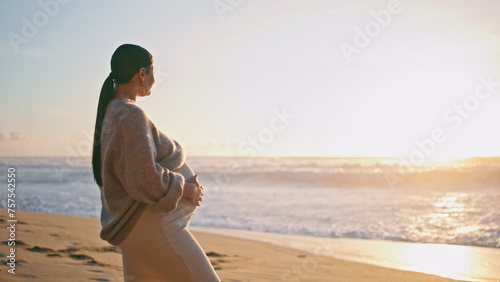 Pregnant woman looking sunset standing on sandy beach. Girl caressing belly photo