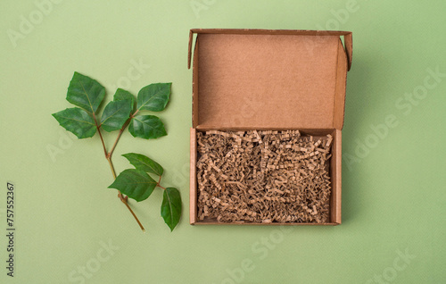 Mockup. Open cardboard box with paper filling on green background with green leaves top view. © Polina Ponomareva