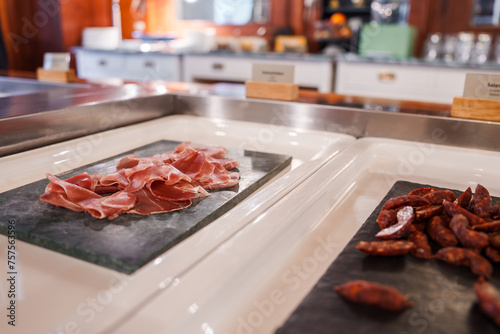 Closeup of a luxury buffet featuring prosciutto on slate trays, with dried sausage and a warm, wooden interior hinting at a Swiss Alps location. Ambient lighting adds to the cozy ambiance.