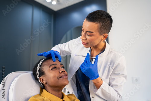 Smiling African woman is getting a rejuvenating facial injections at beauty clinic. The expert beautician is filling female wrinkles by botulinum. Cosmetology skin care.