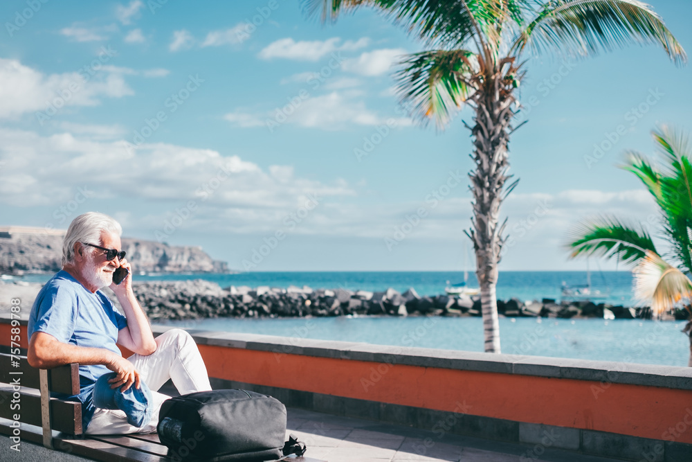 Fototapeta premium Senior bearded man holding smartphone sitting outdoors on bench face the sea enjoying sunny day, travel, vacation, retirement lifestyle