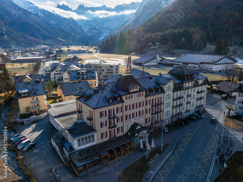 Aerial view of Engelberg resort with the prominent HOTEL BELLEVUE and its redwhite facade. Nearby, clean streets and mountainstyle buildings nestle against a backdrop of majestic, snowcapped peaks. photo