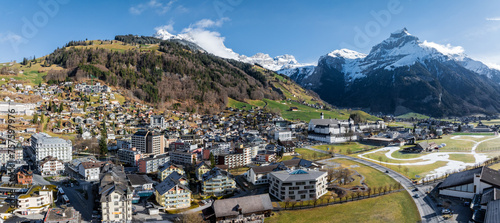 Panoramic view of Engelberg ski resort in Switzerland, showcasing a blend of chaletstyle and modern buildings amid green fields, residual snow, and towering alpine mountains. photo