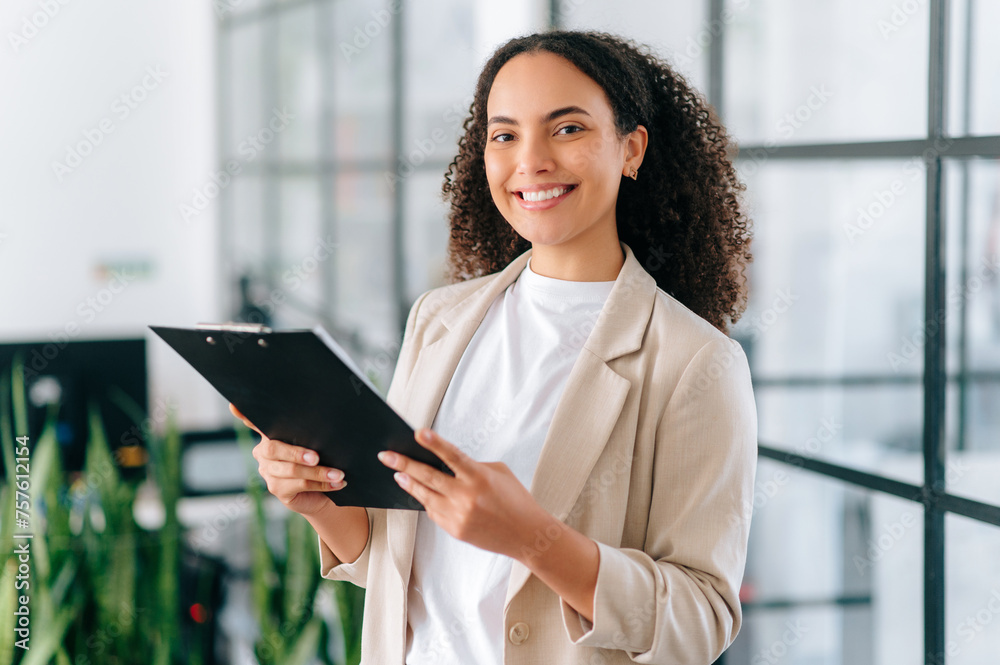 Successful elegant pretty multiracial woman, successful employee of a company, female teacher, standing with documents folder in hands in a modern creative office, looks at camera, smiles friendly Successful elegant pretty multiracial woman, successful employee of a company, female teacher, standing with documents folder in hands in a modern creative office, looks at camera, smiles friendly