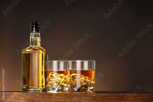 Whiskey with ice cubes in glasses and bottle on wooden table against brown background, space for text © New Africa